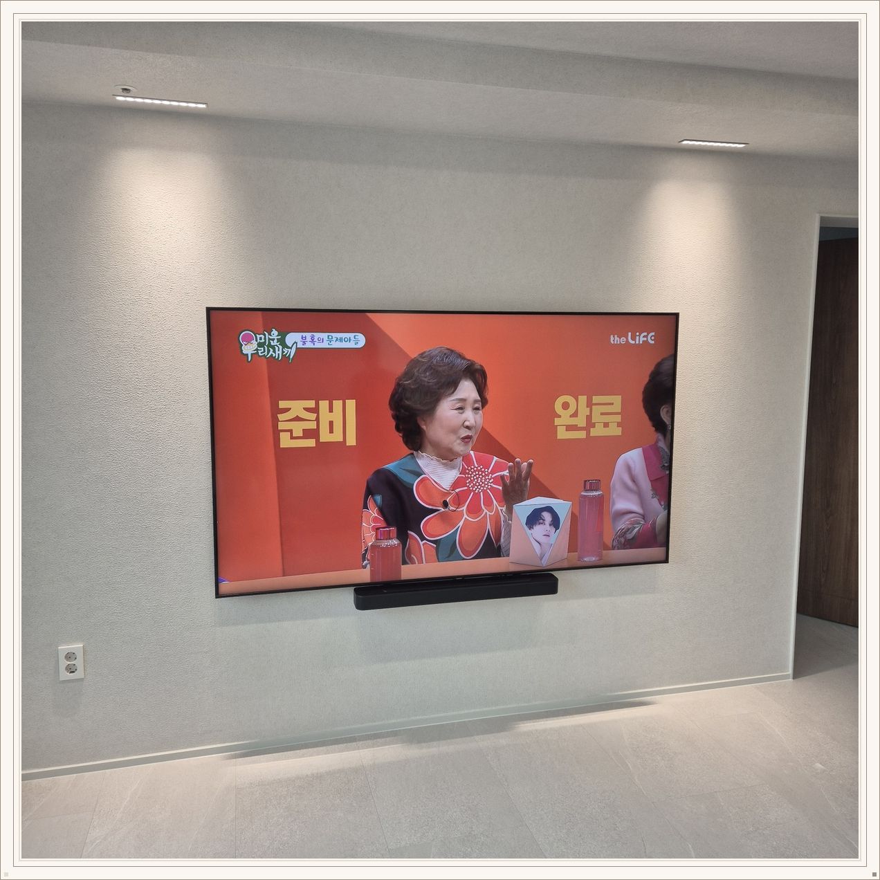 조명이 비추는 미색 벽과 사운드바, 화면 아래 가로로 놓인 검은 음향기기가 선명합니다