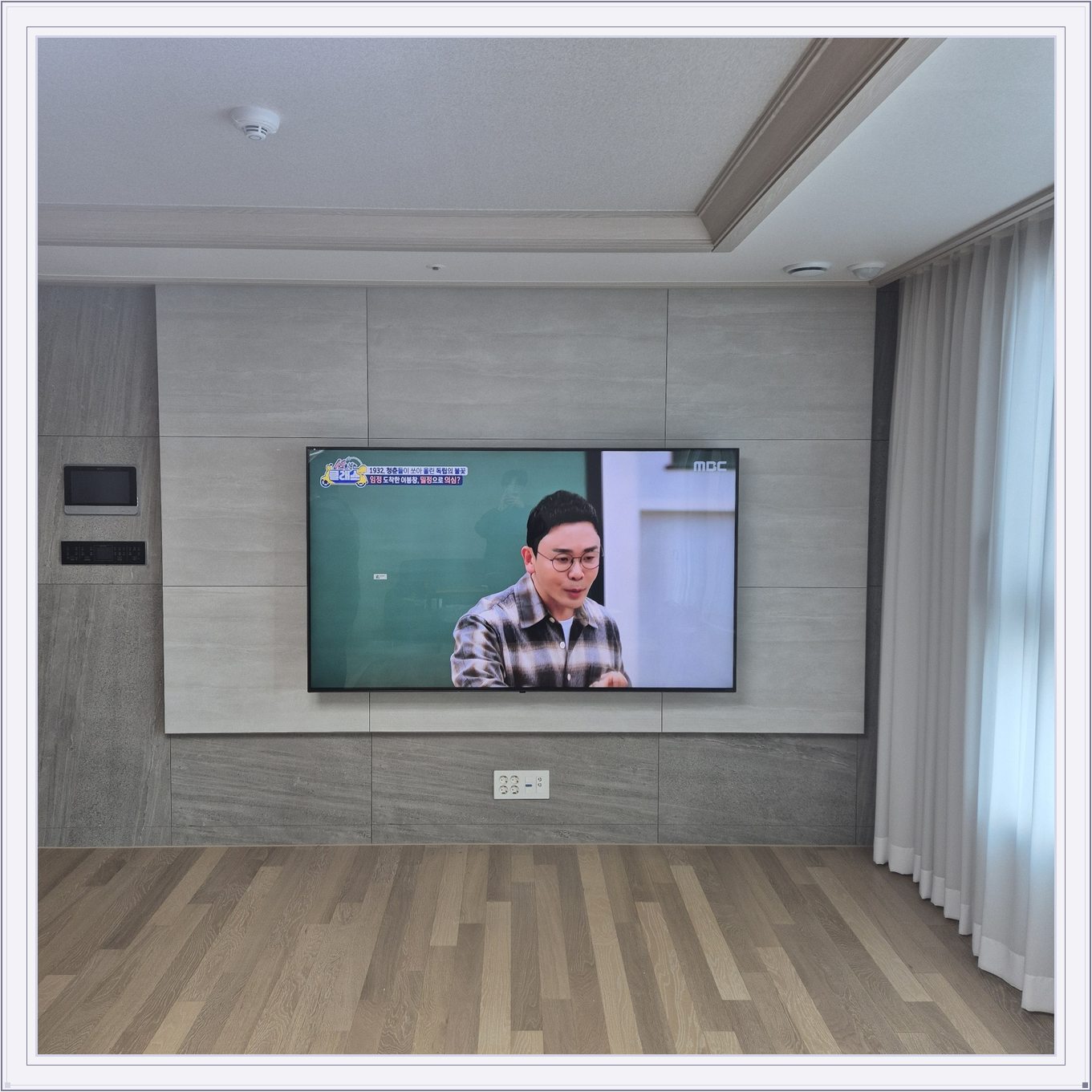 거실 회색 패널과 벽걸이 TV, 오른쪽 커튼 옆으로 화면이 중심에 놓여 있습니다