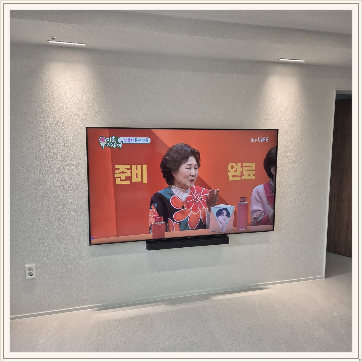 조명이 비추는 미색 벽과 사운드바, 화면 아래 가로로 놓인 검은 음향기기가 선명합니다