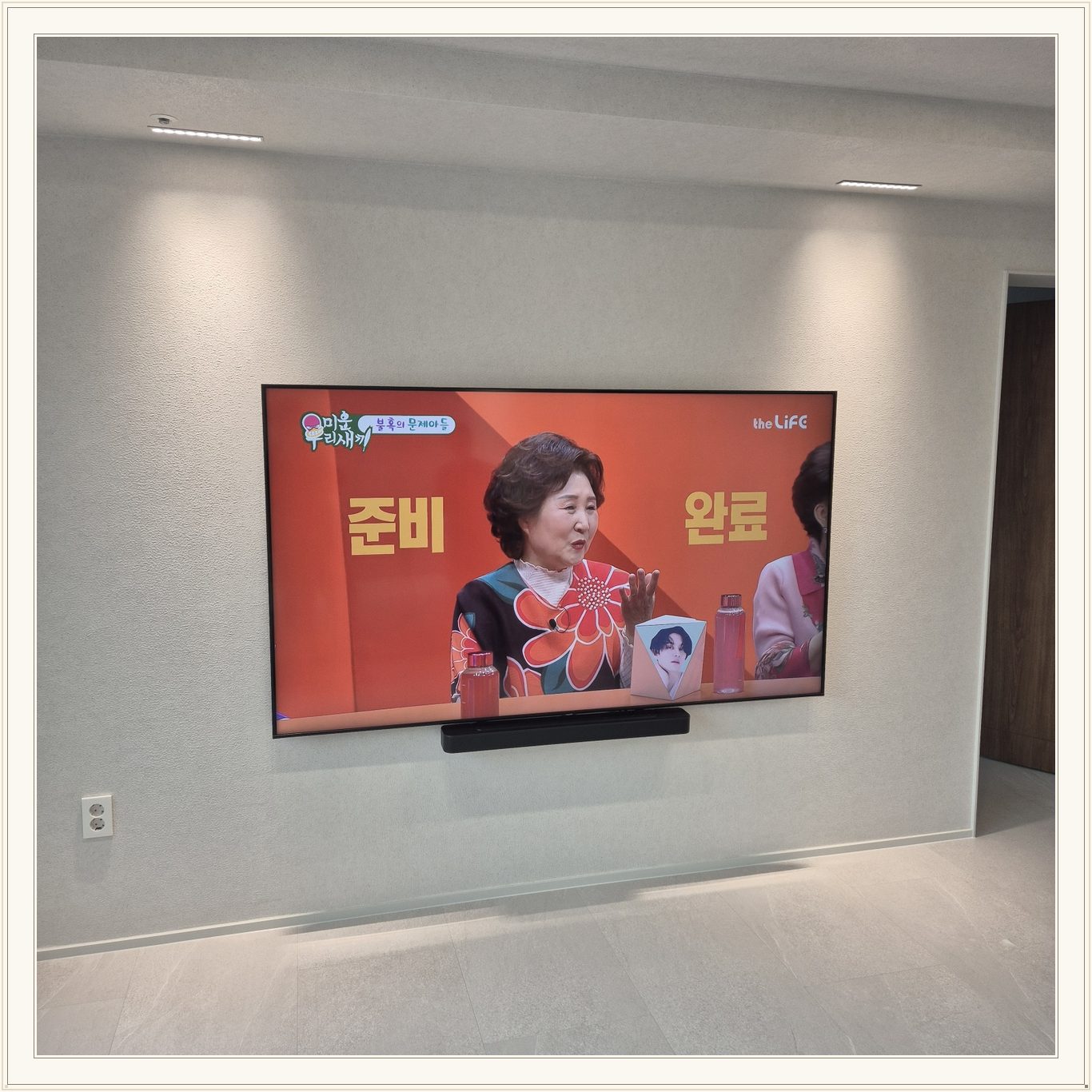조명이 비추는 미색 벽과 사운드바, 화면 아래 가로로 놓인 검은 음향기기가 선명합니다