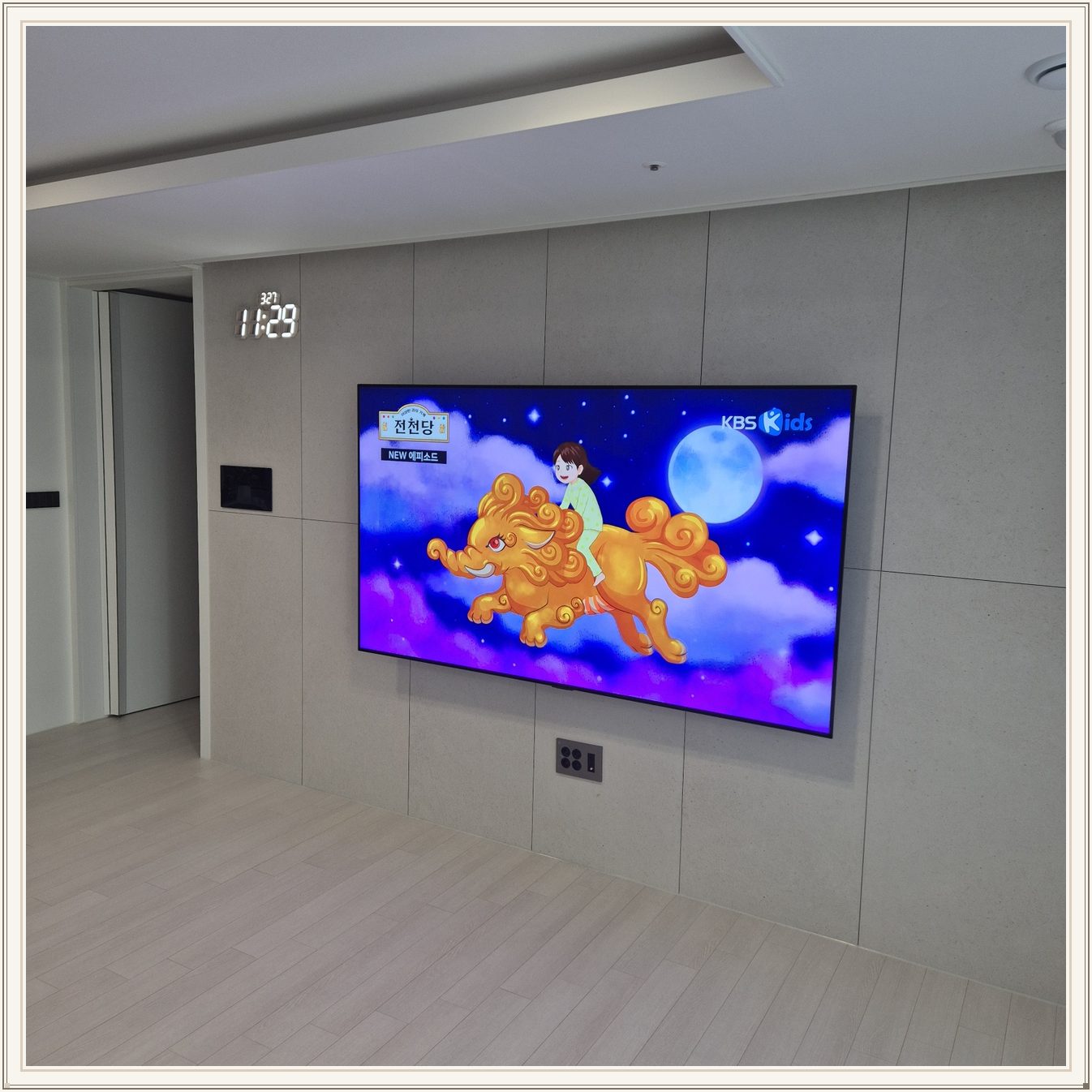 회색 판넬 벽 위의 화면과 디지털 시계, 벽면 패널 이음선과 TV 중심의 배치를 비교할 수 있습니다