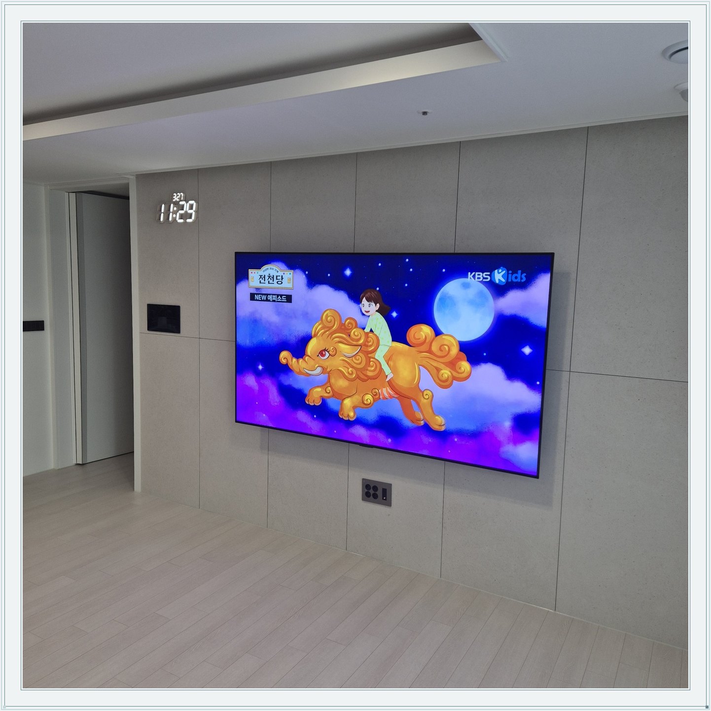 회색 판넬 벽 위의 화면과 디지털 시계, 벽면 패널 이음선과 TV 중심의 배치를 비교할 수 있습니다