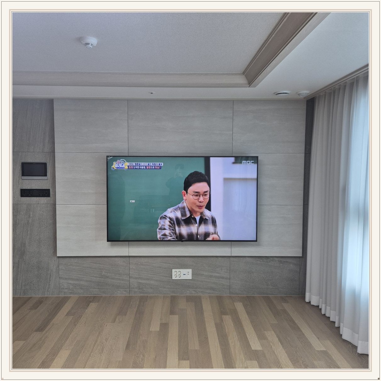 거실 회색 패널과 벽걸이 TV, 오른쪽 커튼 옆으로 화면이 중심에 놓여 있습니다