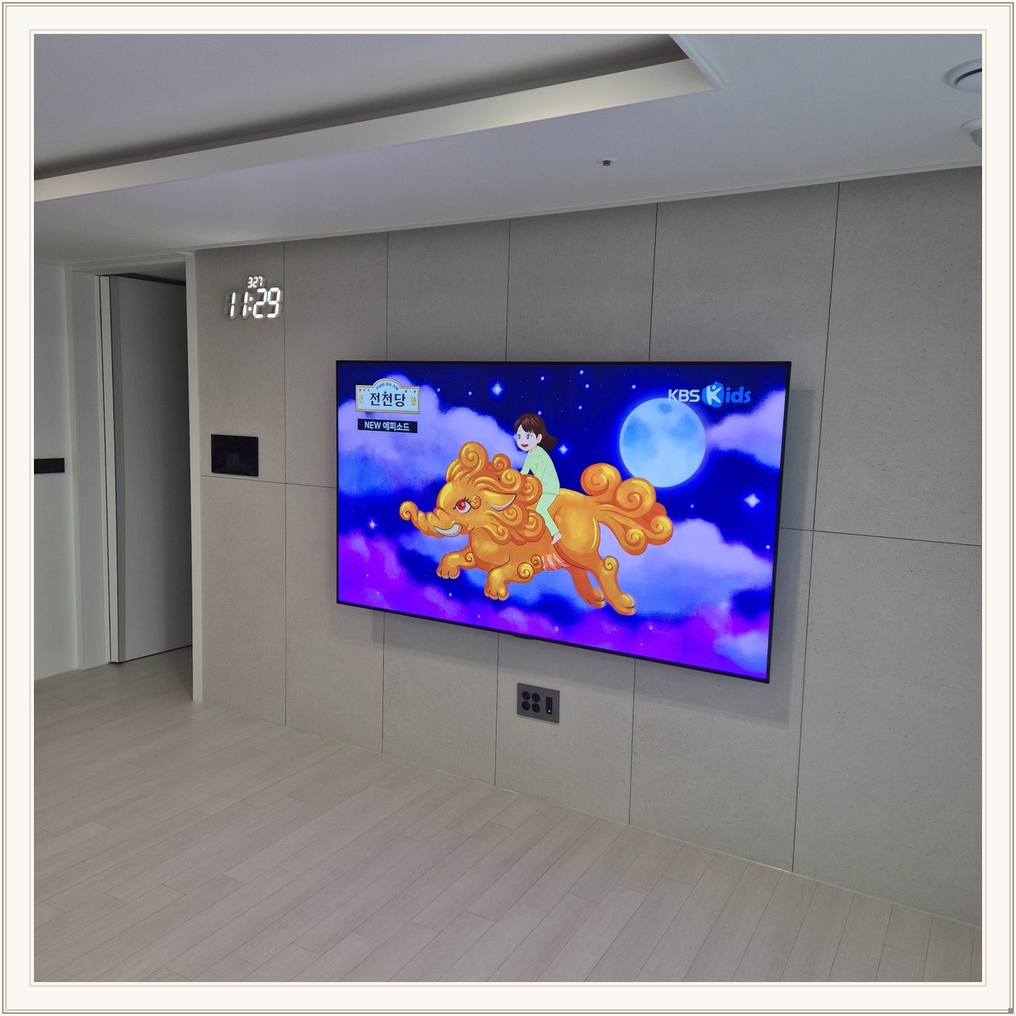 회색 판넬 벽 위의 화면과 디지털 시계, 벽면 패널 이음선과 TV 중심의 배치를 비교할 수 있습니다
